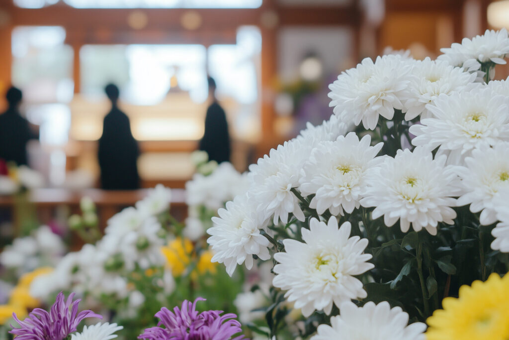祭壇前で参列者が静かに佇む葬儀会場の様子と、手前に並ぶ白い菊の花