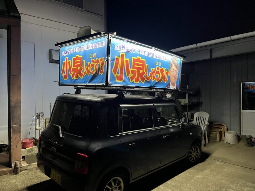 夜間に撮影されたスズキ・ラパンの選挙カー。屋根の上に大型の発光看板を載せ、「小泉しゅうすけ」と書かれた選挙用ボードが明るく点灯している。