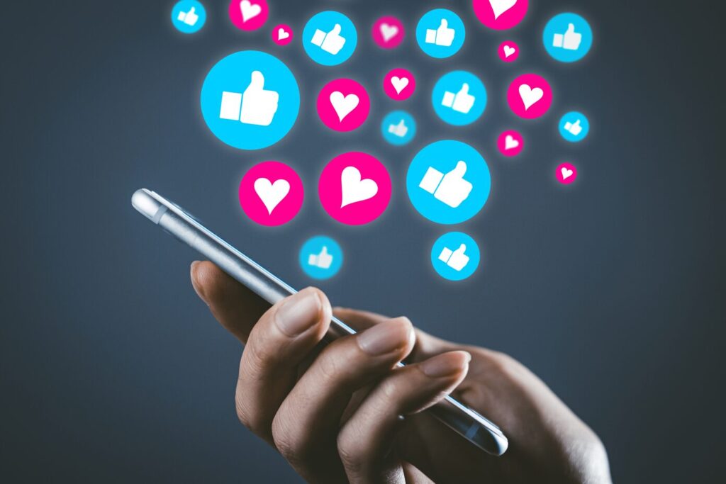 スマートフォンを操作する手の上に、SNSの「いいね」やハートマークが浮かび上がるイメージ画像。SNS利用やオンラインコミュニケーションを象徴するビジュアル。